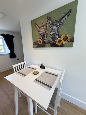 A dining area with a white table, two chairs, a painting of horses and sunflowers, and a window with curtains. - The Traveller`s Loft, The Crown Inn (Cerney Wick)