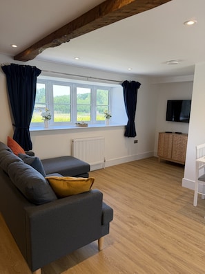 A modern living room with a grey sofa, wooden flooring, a radiator, a TV, and a window with curtains. - The Traveller`s Loft, The Crown Inn (Cerney Wick)
