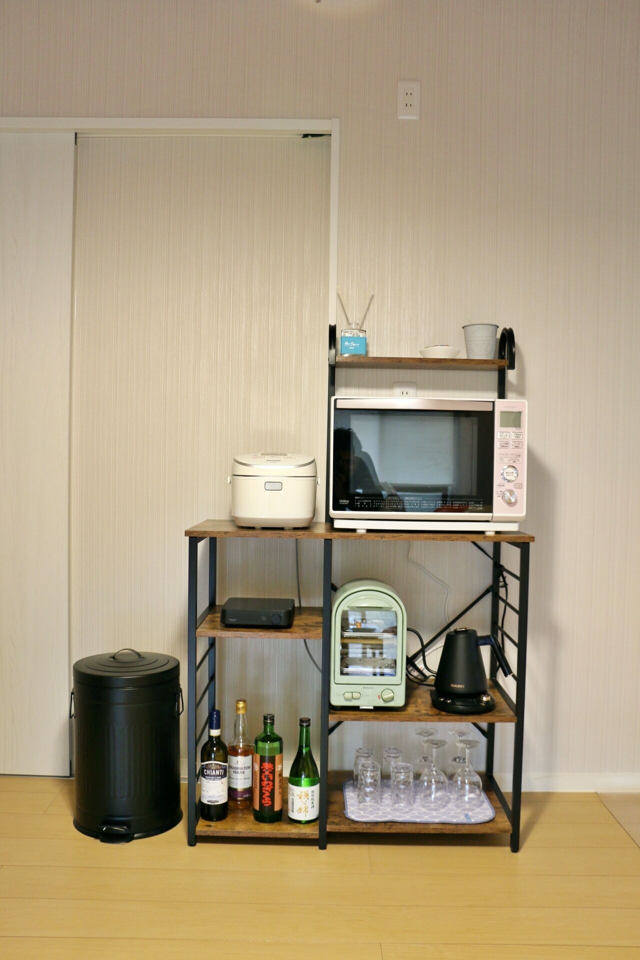 A kitchenette with a microwave, rice cooker, and a shelf with bottles and glasses.