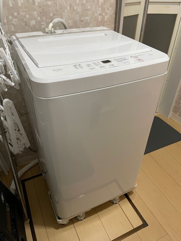 A white, portable washing machine with a control panel, placed on a wooden floor in a room with a tiled wall.