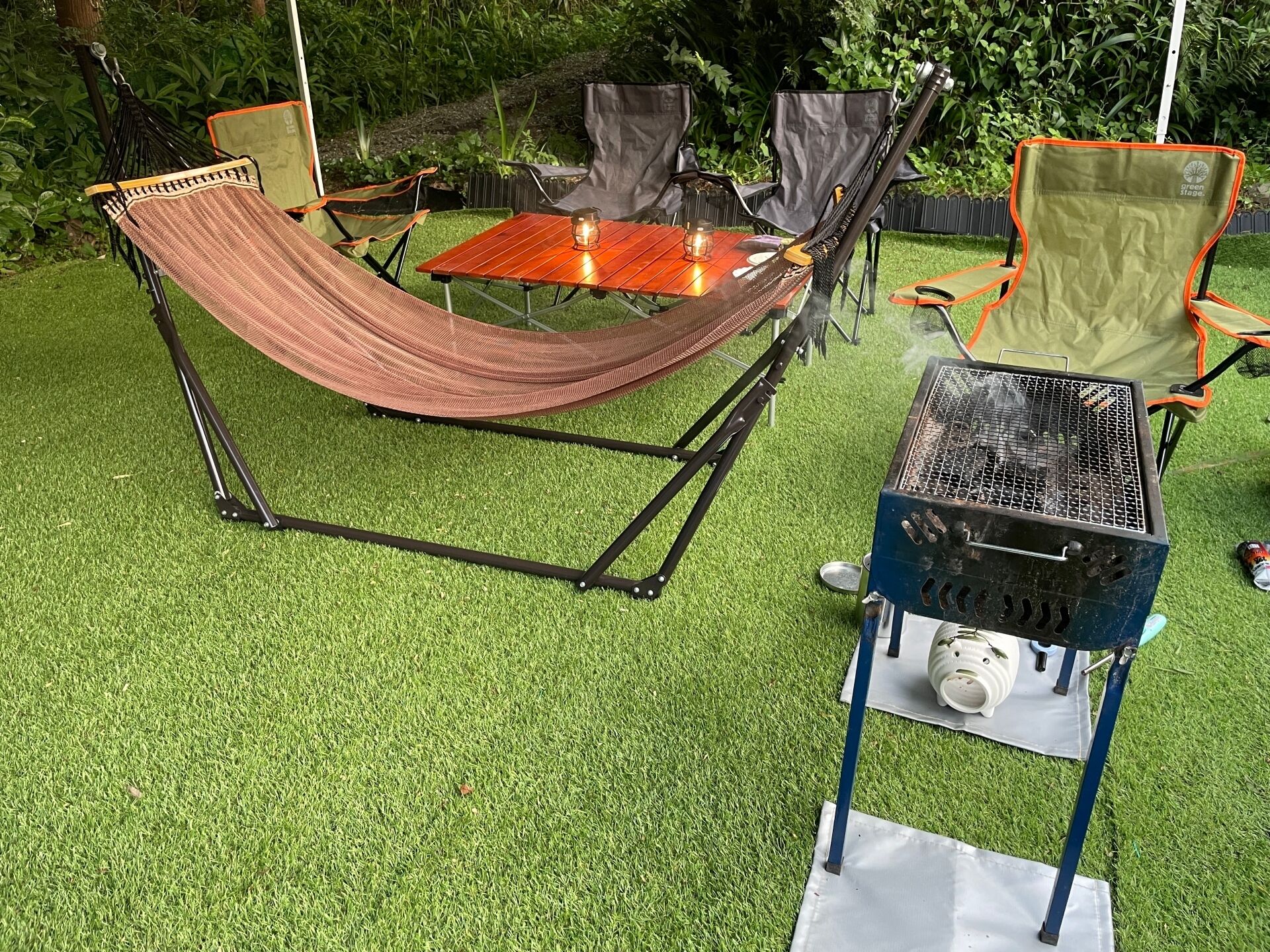 A hammock set up over a table with a grill and chairs in a grassy outdoor area.