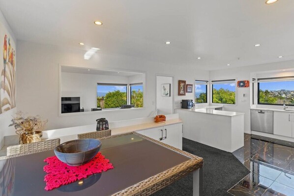A modern kitchen with white cabinetry, a dining area with a black table, and large windows with a view of greenery.