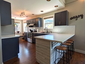 A modern kitchen with dark cabinets, a large island with stools, and a 'enjoy' sign on the wall. - Kimberley Mountain Getaway | Ski, Golf & Adventure (Kimberley)