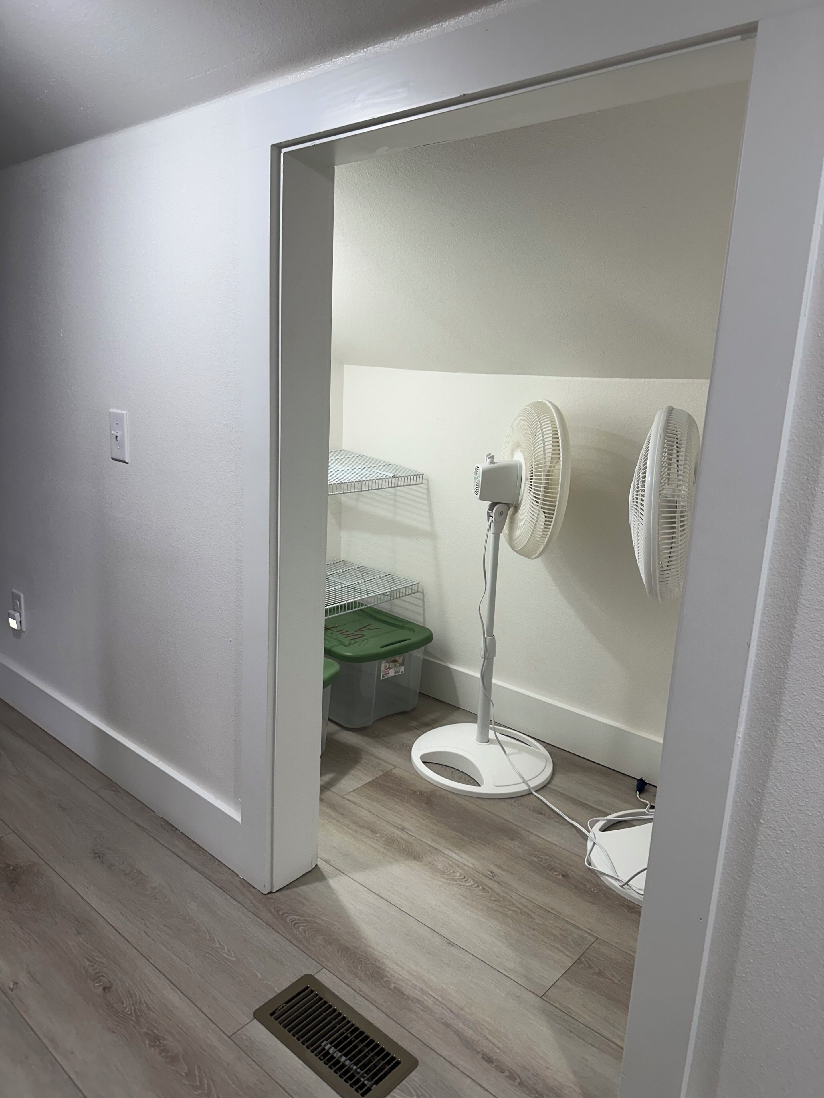 Ein Zimmer mit weißer Wand, weißem Boden, einem weißen Standventilator, einer grünen Aufbewahrungsbox und einer weißen Steckdose.