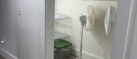 Ein Zimmer mit weißer Wand, weißem Boden, einem weißen Standventilator, einer grünen Aufbewahrungsbox und einer weißen Steckdose.