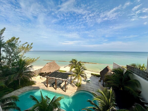 A pool area with a view of the ocean, a thatched-roof structure, and palm trees.