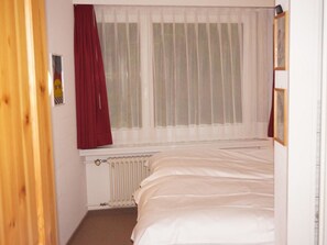 A bedroom with a bed, a radiator, a window with sheer curtains, and a partial view of a door. - Paradies 104 Appartement/Fewo, Dusche und Bad, WC, 4-Bettwohnung 4-Bettwohnung by Interhome (Arosa)