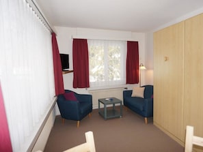 Une petite pièce bien éclairée, avec deux fauteuils bleus, une petite table, un téléviseur posé sur un meuble, un radiateur et une armoire en bois.