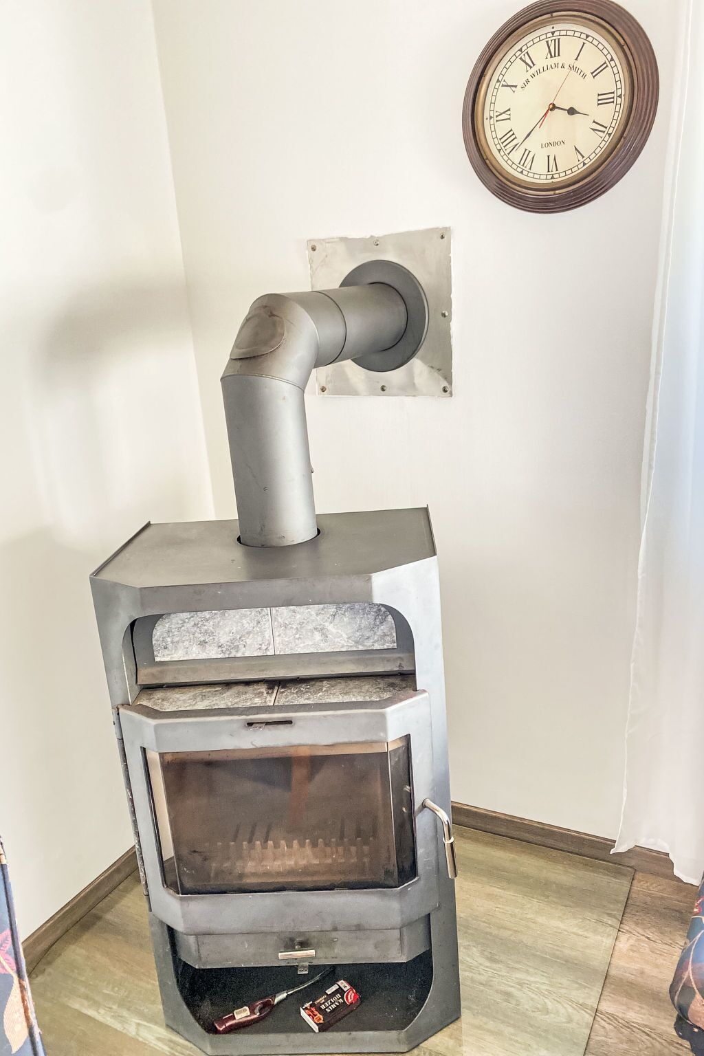 A wood stove with a chimney, a clock on the wall, and a small box of tissues on the floor.