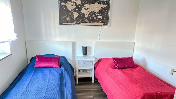 Una habitación pequeña y acogedora con dos camas individuales, cada una con almohada y manta. Hay una mesita de noche blanca pequeña con una lámpara entre las camas. Un mapa del mundo cuelga en la pared sobre las camas.