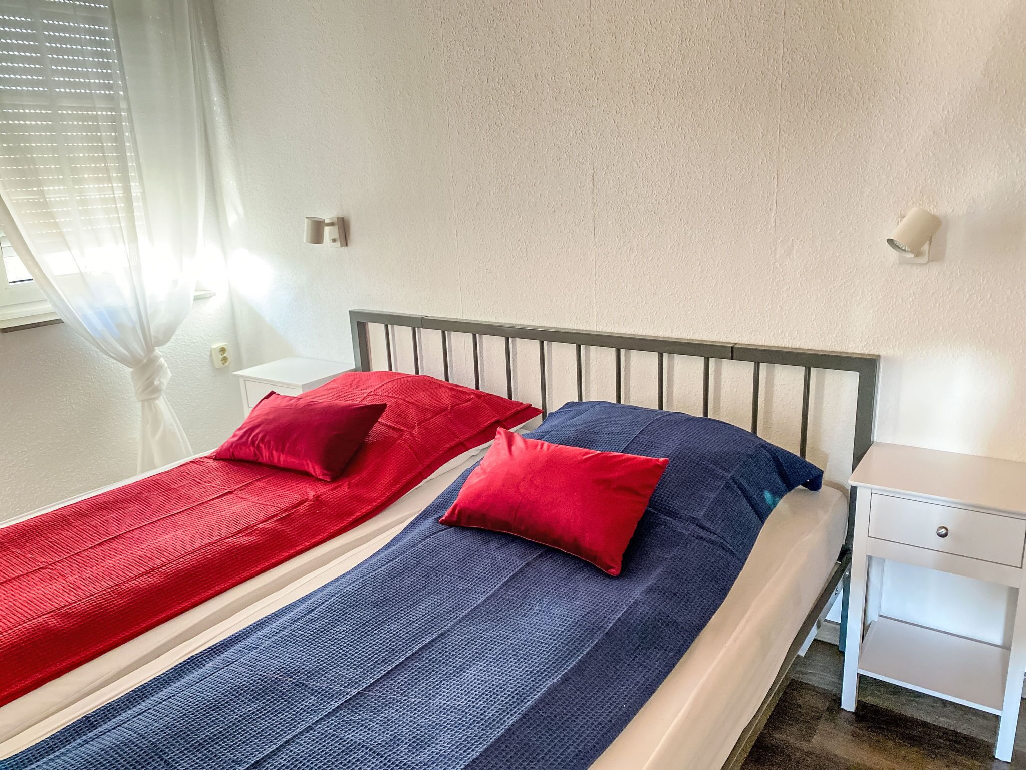 A single bed with a metal headboard, two pillows, a white nightstand, and a window with blinds.