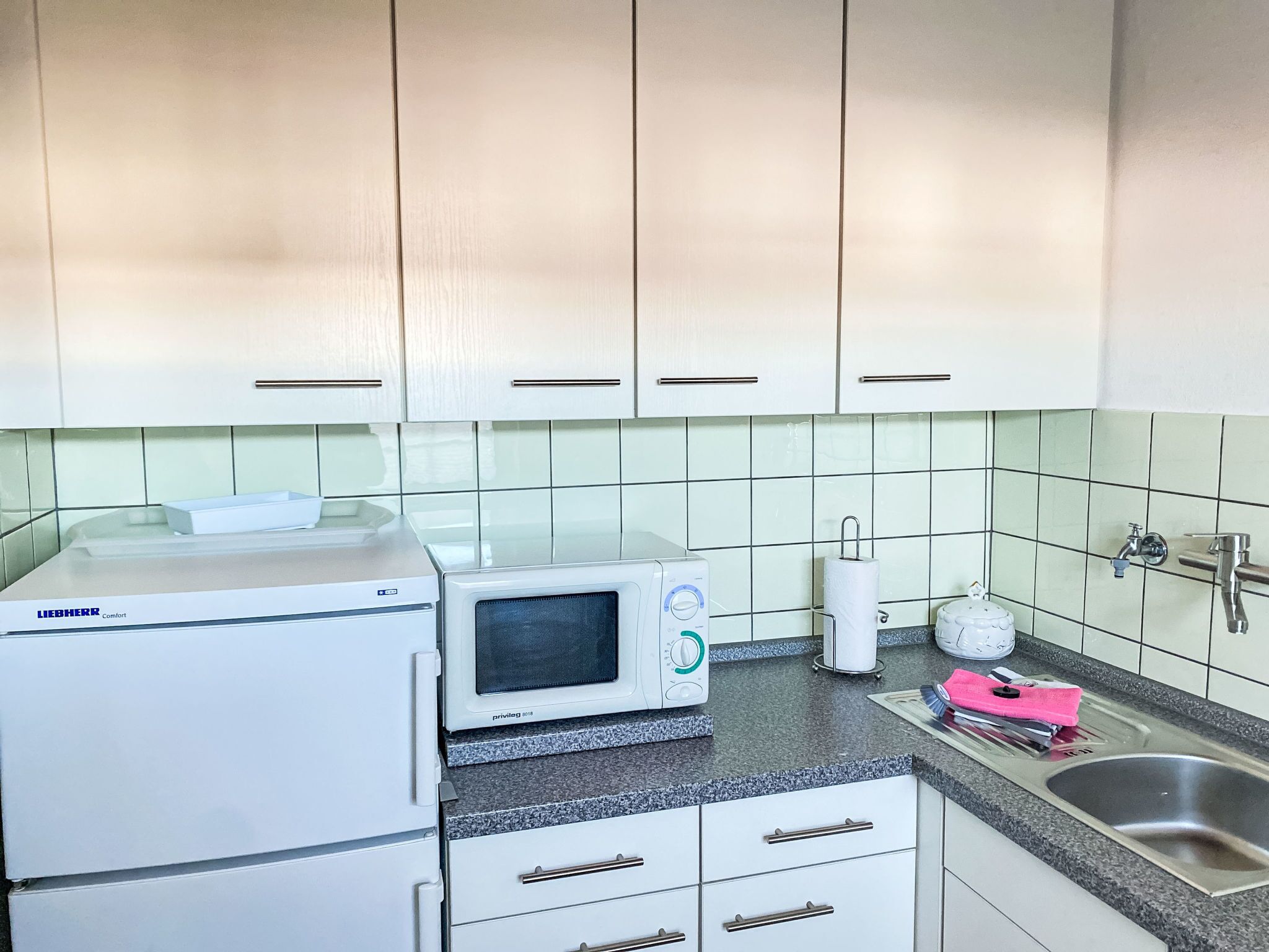 A kitchen with white cabinets, a Liebherr Comfort refrigerator, a microwave, a sink, and a tiled backsplash.