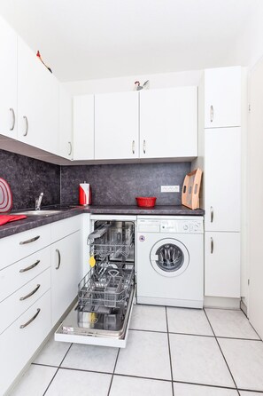 A compact kitchen with white cabinets, a built-in dishwasher, and a countertop with a red basket. - Jupiter by Interhome (Dornumersiel)