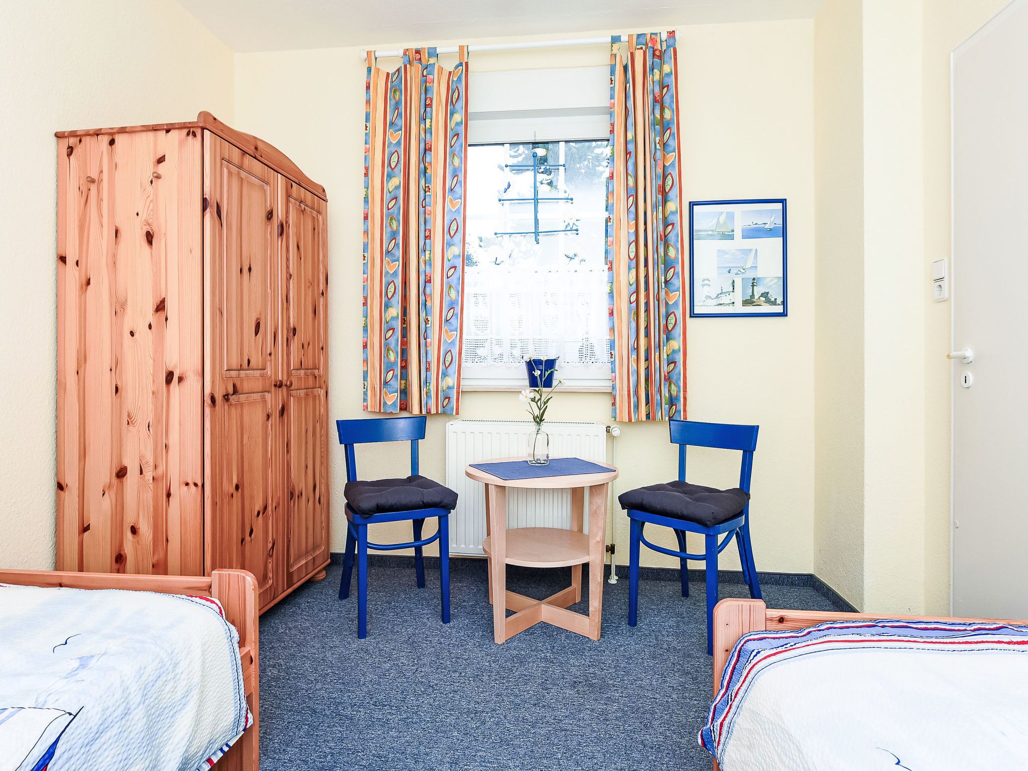 A room with two single beds, a wooden wardrobe, a small table with a chair, and a window with curtains.
