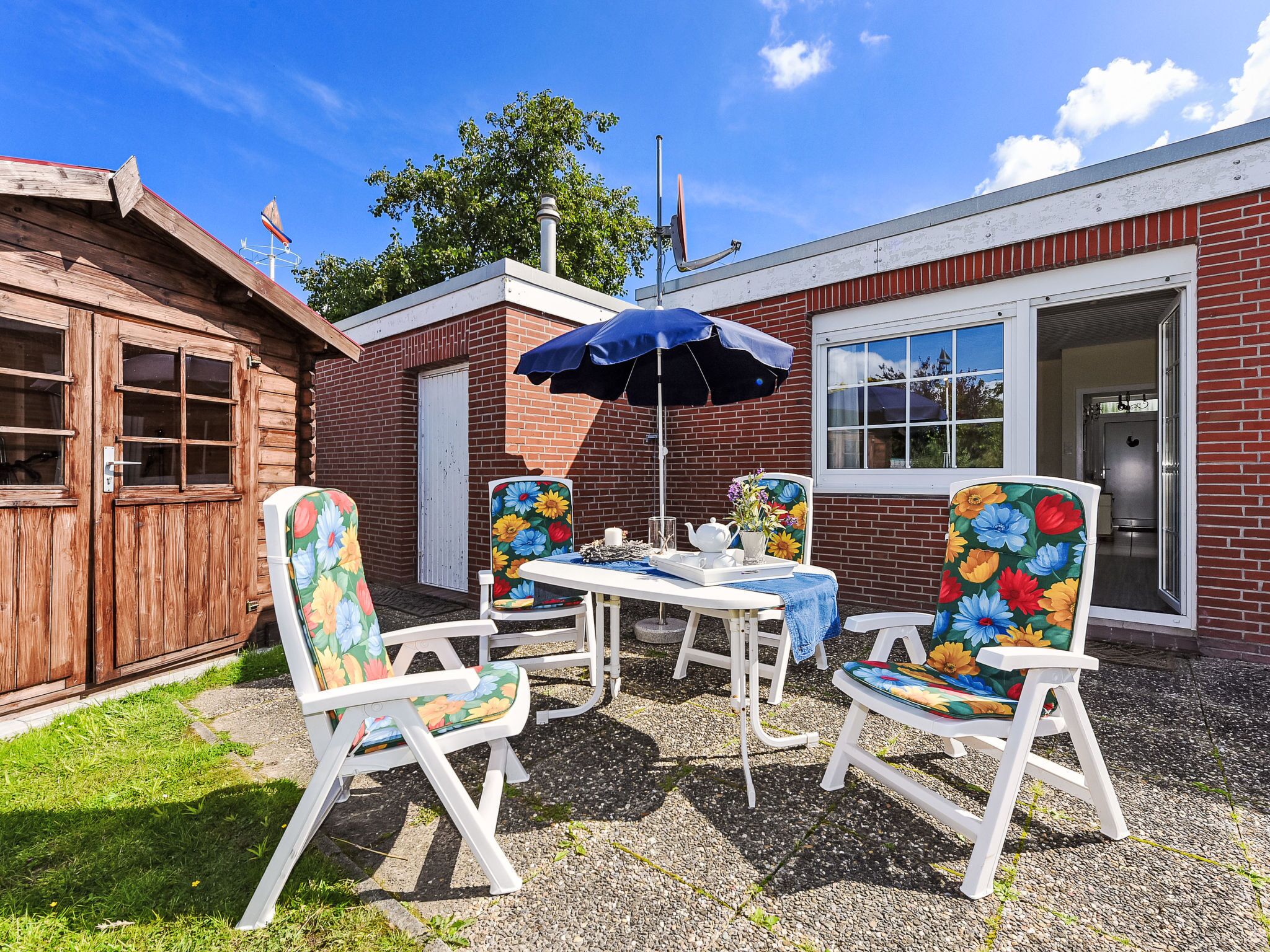 An outdoor patio with a table and four chairs, a blue umbrella, and a brick building with a wooden door.