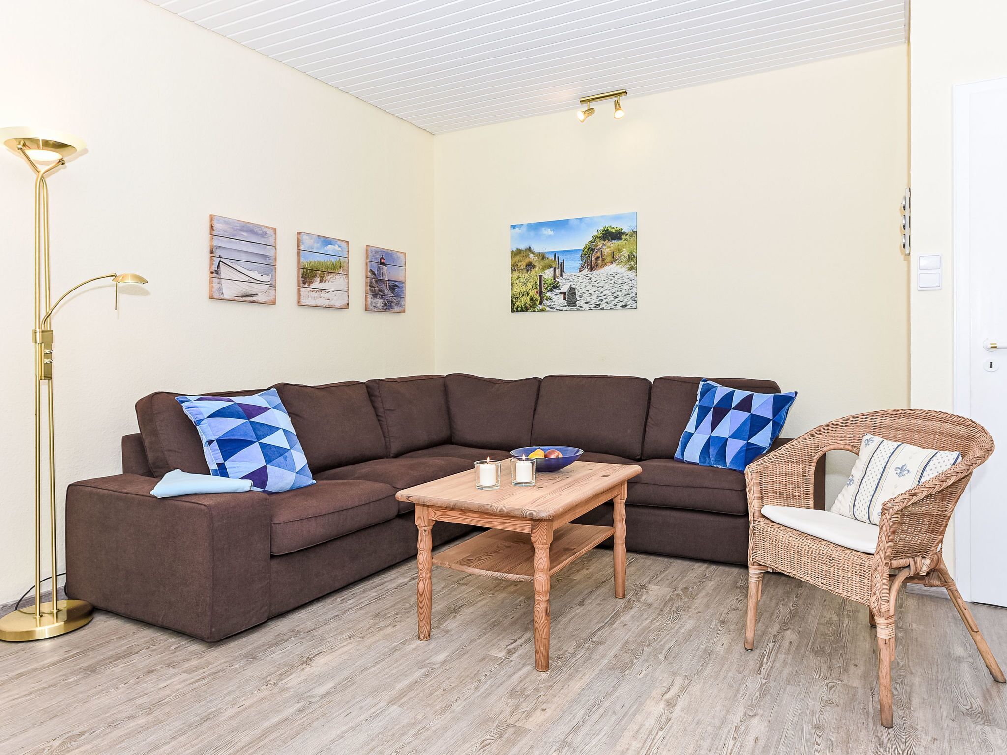 A living room with a brown sectional sofa, a wicker chair, a wooden coffee table, and wall art depicting coastal scenes.
