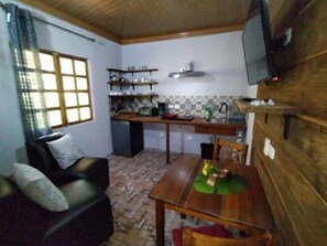 A compact living space with a dining table, a sofa, a television mounted on the wall, and a kitchen area with shelves and a stove.