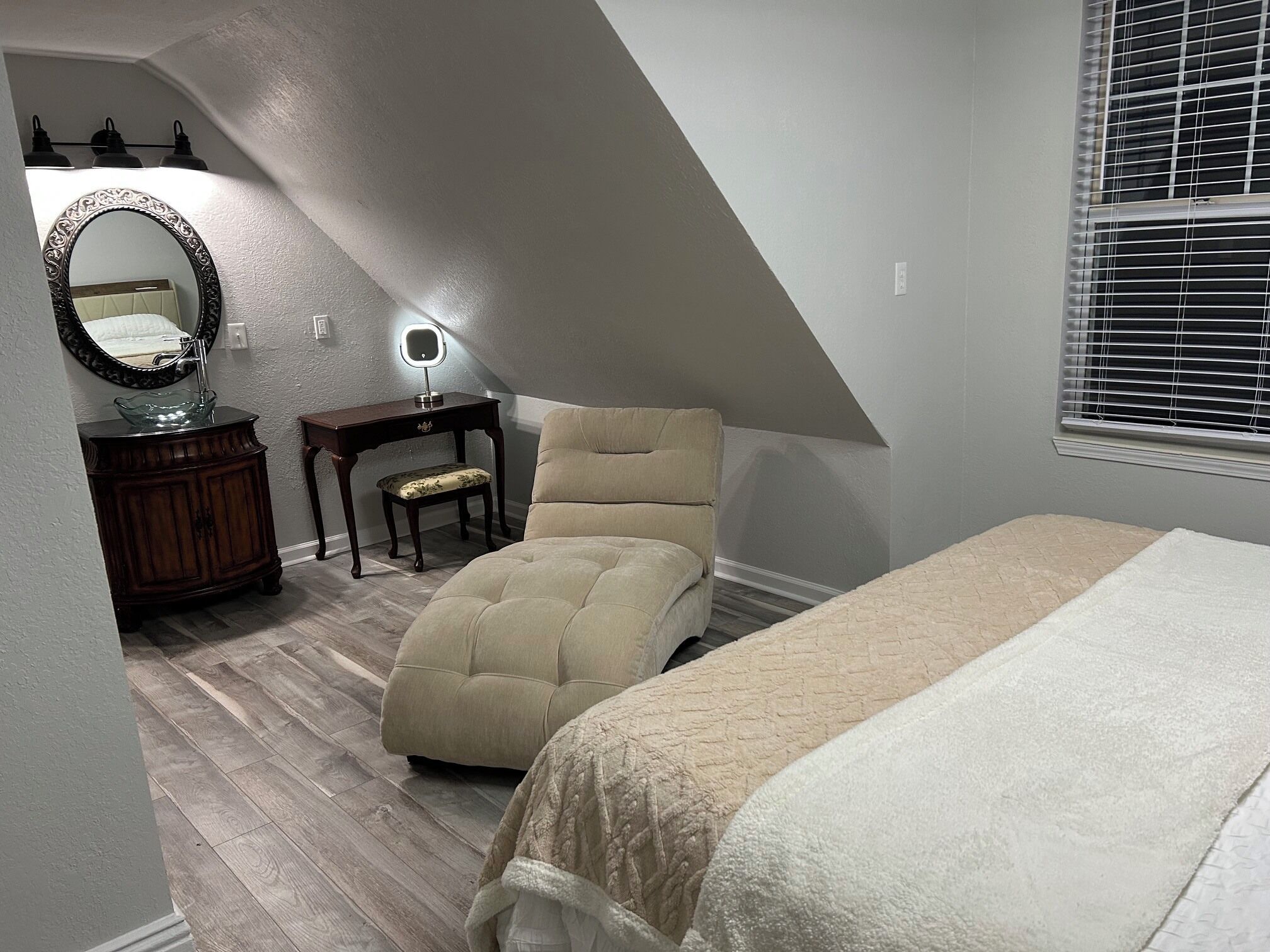 A bedroom with a sloped ceiling, a bed, a chair, a vanity with a mirror, and a window with blinds.