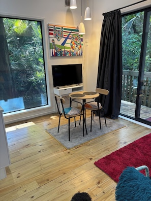 A modern living room with a flat-screen TV, a dining table, two chairs, a red rug, and a large window with a view of greenery.