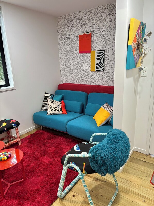 A modern living room with a blue sofa, red and yellow cushions, a red shag rug, a blue furry chair, and a wall with abstract art.