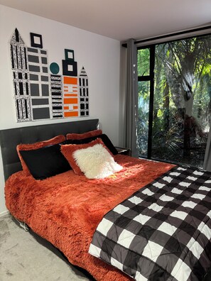 A bedroom with a large bed, a cityscape wall art, and a view of greenery through the window. - Heritage reserve hideaway.Bush all around. One Bed-ground floor of our home (Auckland)
