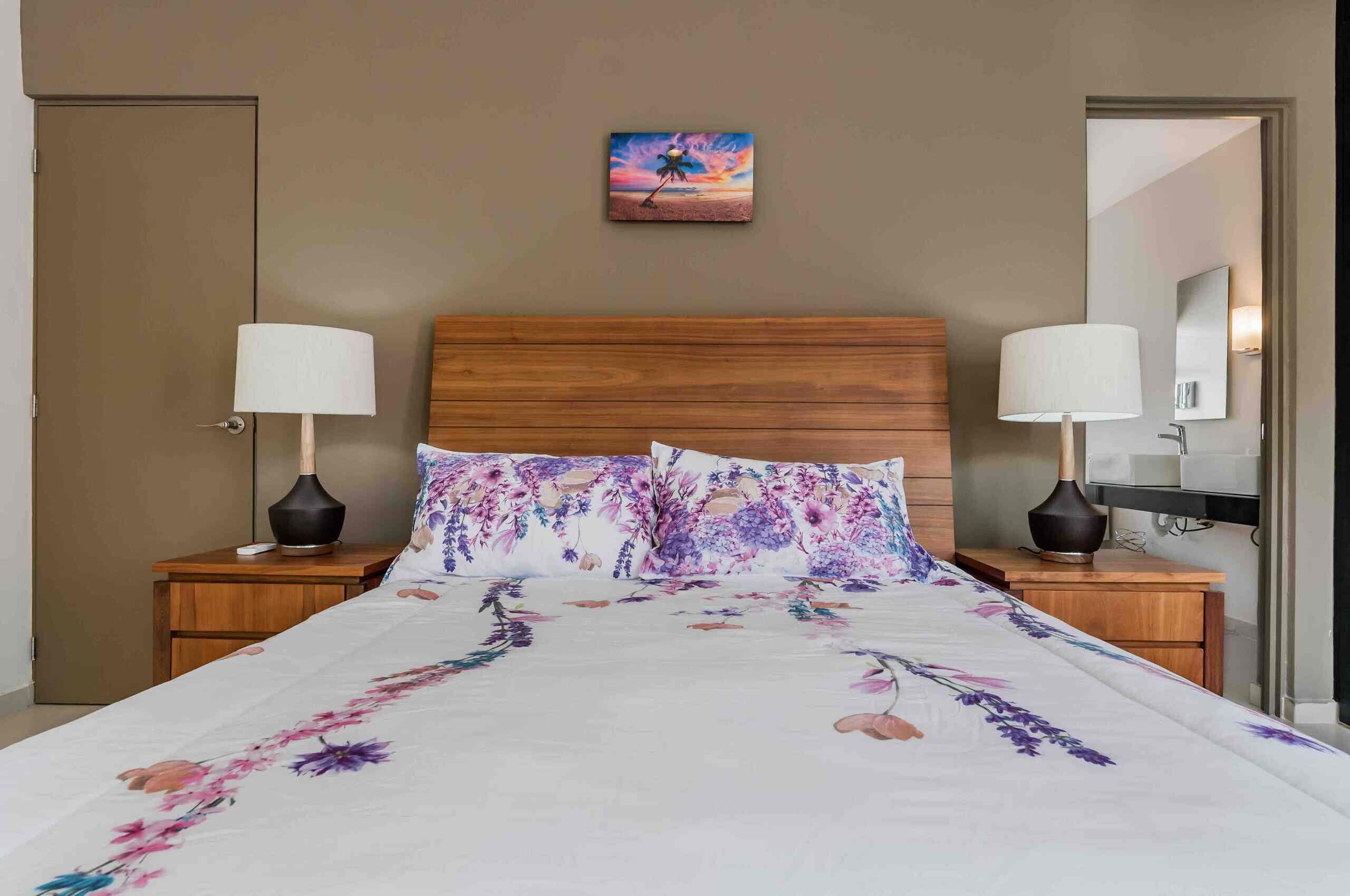 Une chambre d’hôtel avec un grand lit, une literie à motifs floraux, deux tables de chevet avec des lampes, une tête de lit en bois et un tableau au mur.