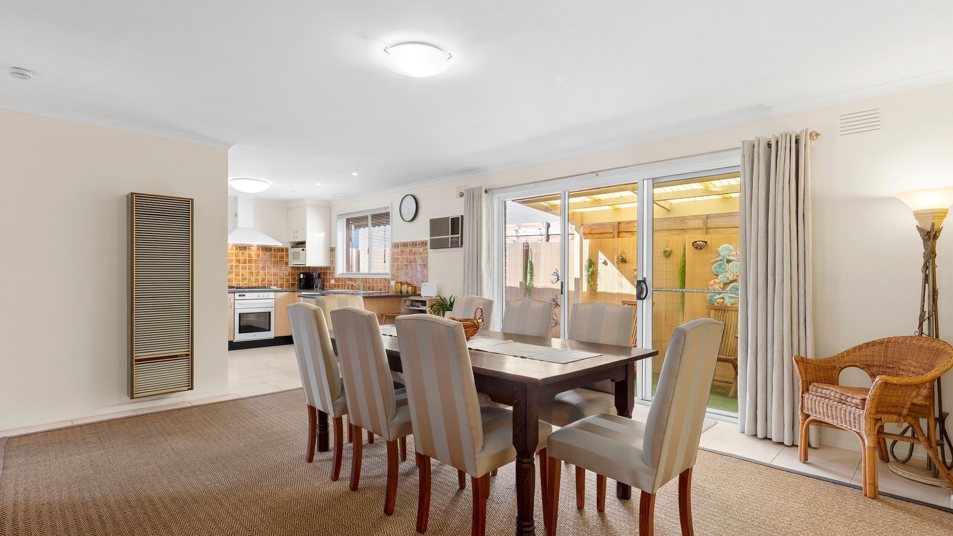 A dining area with a table and chairs, a kitchen in the background, and a view of the outdoors through a glass door.