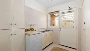A compact kitchen with a built-in washing machine, a sink, and a window with a view of a brick wall. - Acacia - Point Lonsdale (POINT LONSDALE)