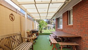 An outdoor patio with wooden benches and picnic tables, a covered area with a roof, and a brick wall. - Acacia - Point Lonsdale (POINT LONSDALE)
