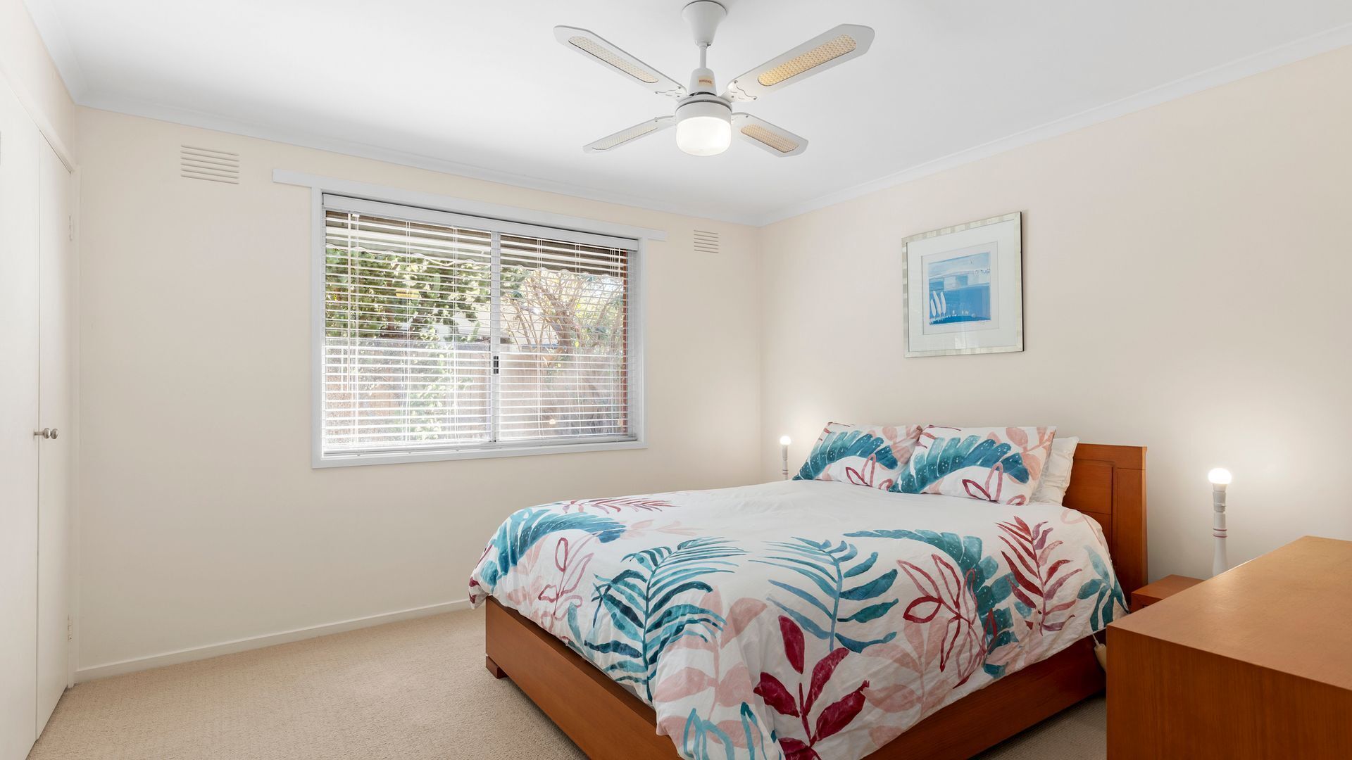 A bedroom with a bed, a ceiling fan, a window with blinds, a bedside table with a lamp, and a framed picture on the wall.