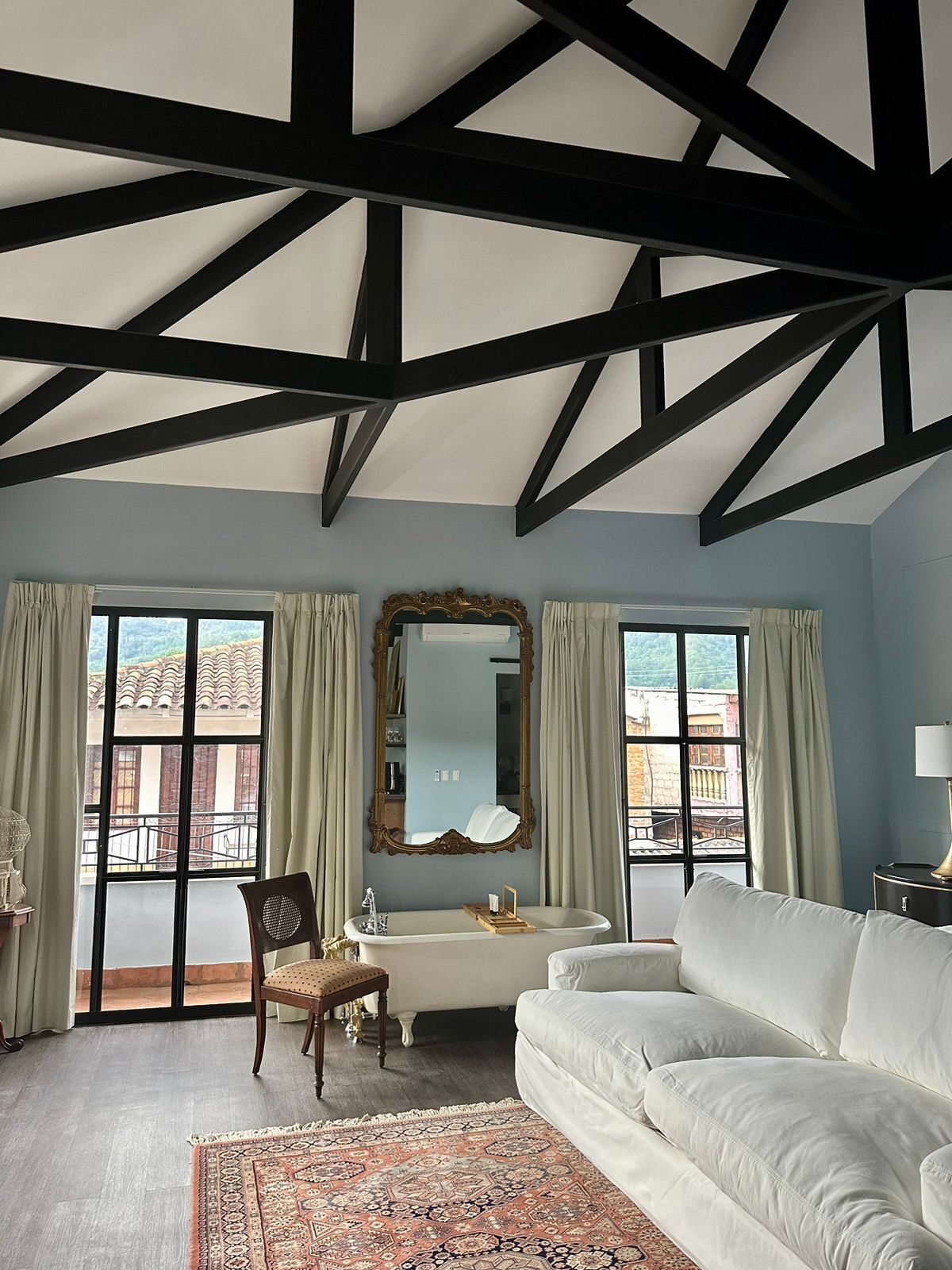 A room with a high ceiling, a white sofa, a large mirror, a bathtub, and a view of buildings through large windows.