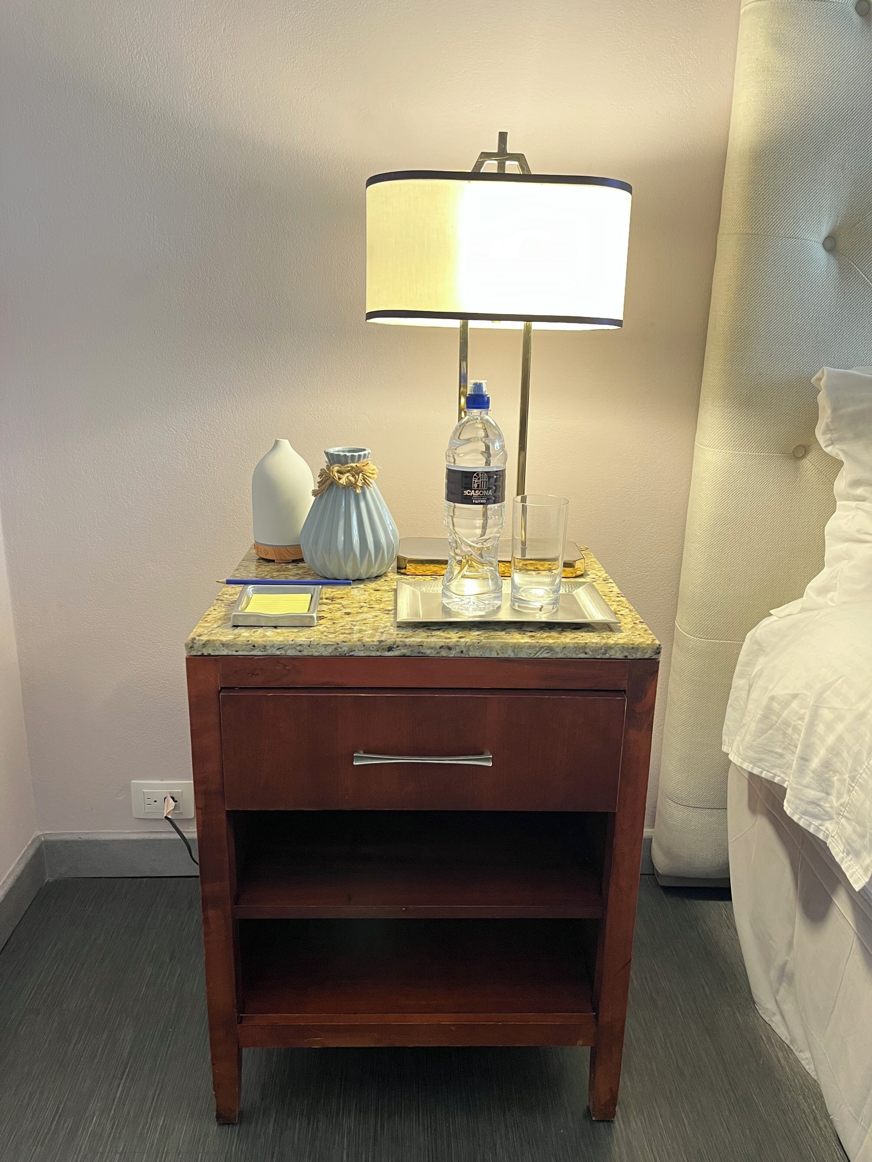 A wooden bedside table with a lamp, a vase, a water bottle, and glasses.