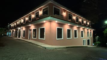 A two-story building with multiple windows and balconies, featuring a flat roof and a paved ground.