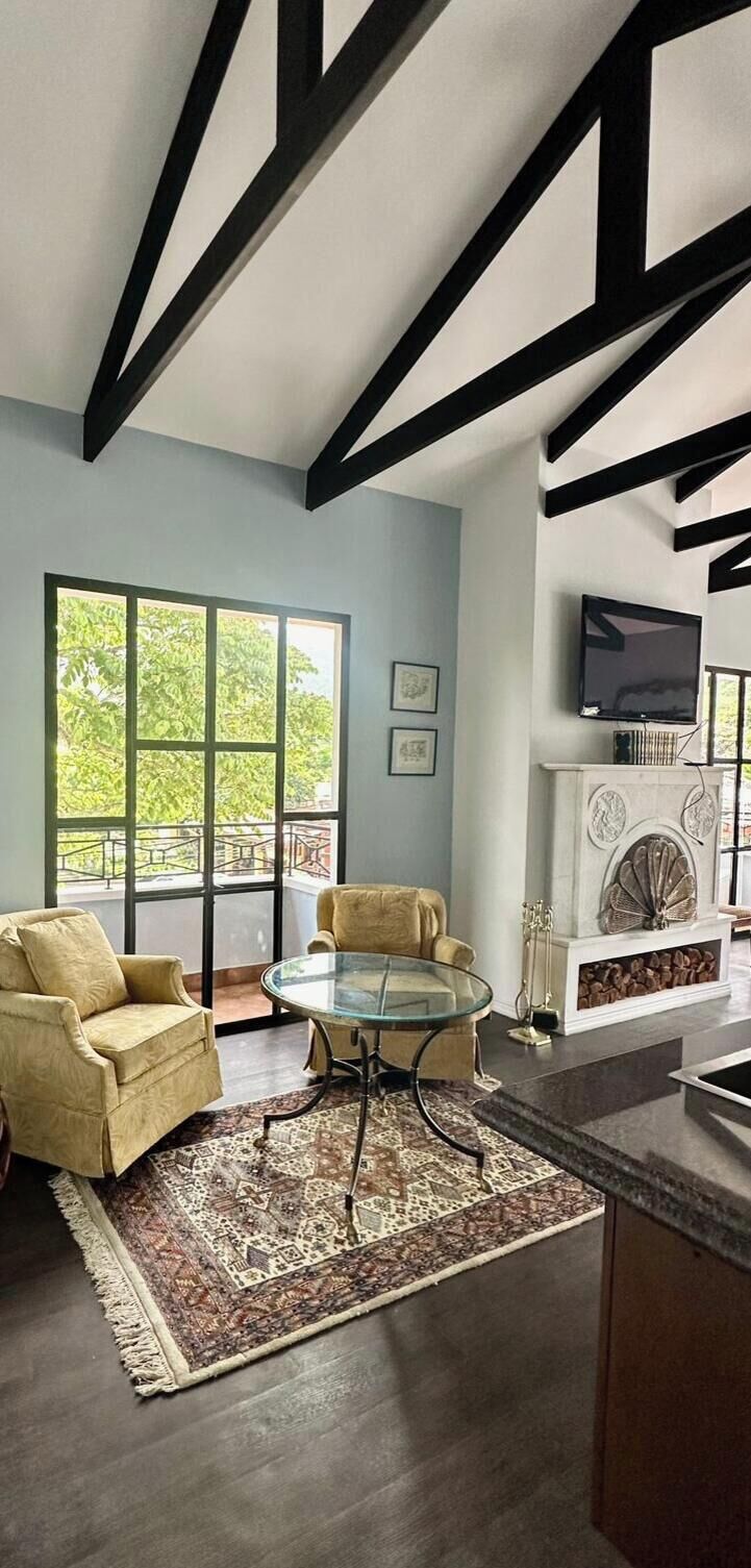 A modern living room with a glass coffee table, two sofas, a fireplace, and a large window with a view of greenery.