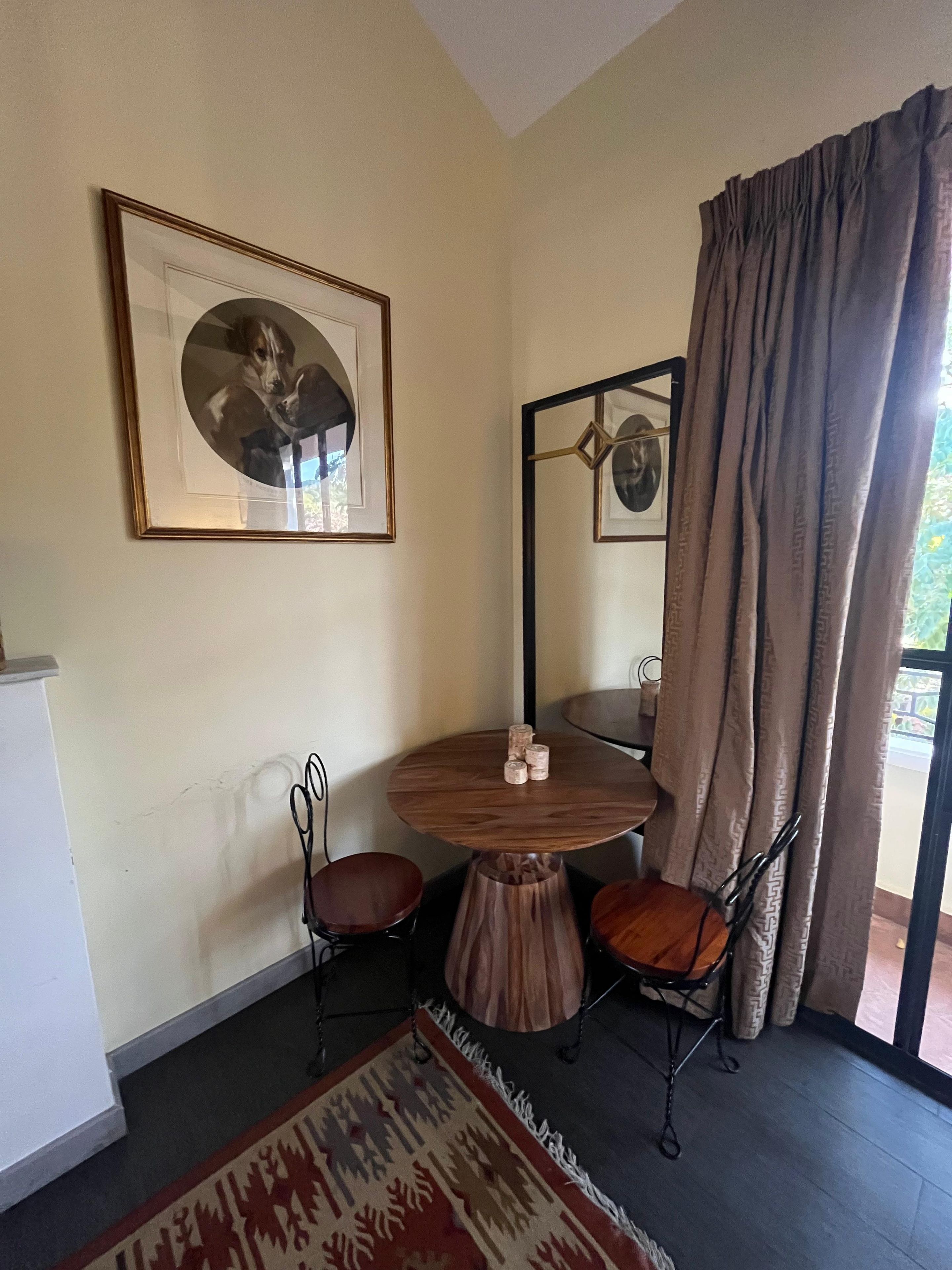 A room with a round wooden table, two chairs, a framed picture on the wall, a mirror, and a window with curtains.