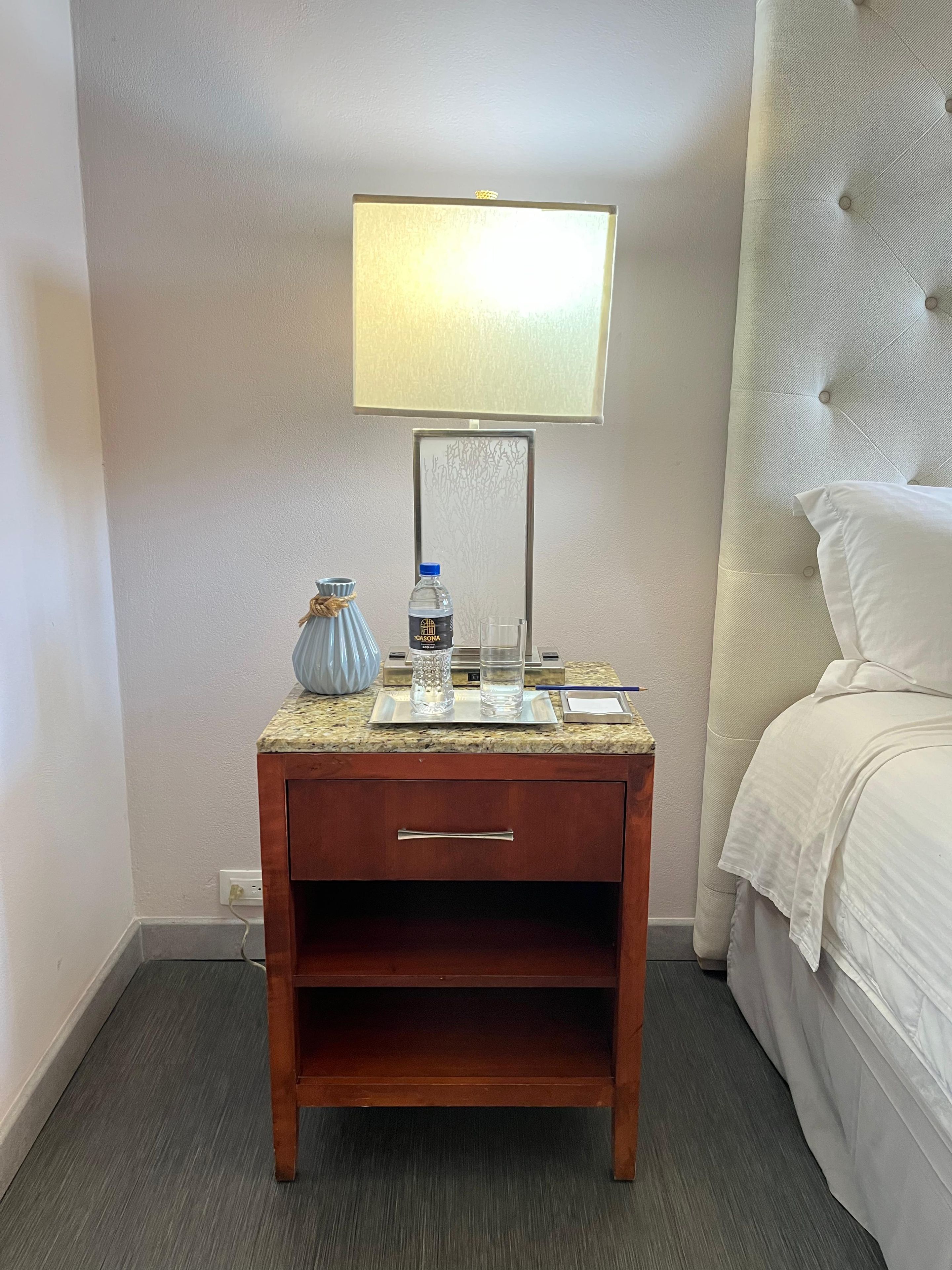 Habitación de hotel con cama, mesita de noche con lámpara, botella de agua, vaso y florero.