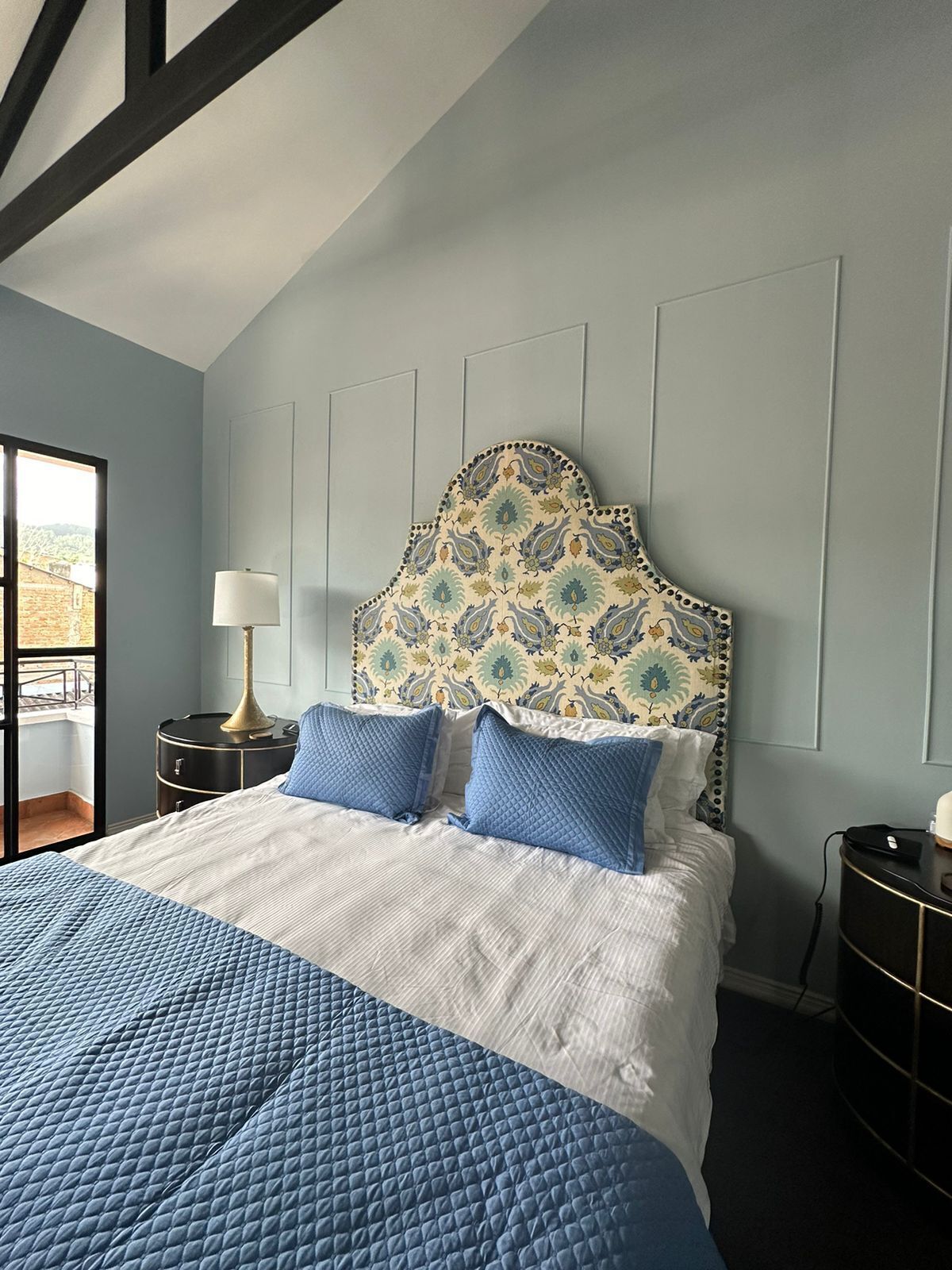 A bedroom with a bed, a nightstand, a lamp, and a view of the outside.