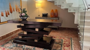 Un moderno comedor con una mesa de madera oscura, una planta en maceta y una lámpara. Se ve una escalera con barandillas de cristal al fondo.