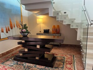 A modern dining area with a dark wooden table, a potted plant, and a lamp. A staircase with glass railings is visible in the background. - Boutique Hotel La Casona  (Valle de Angeles)