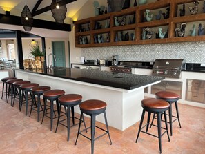 Una cocina moderna con una isla central, taburetes de bar, salpicadero con diseño y un estante de madera con objetos decorativos.