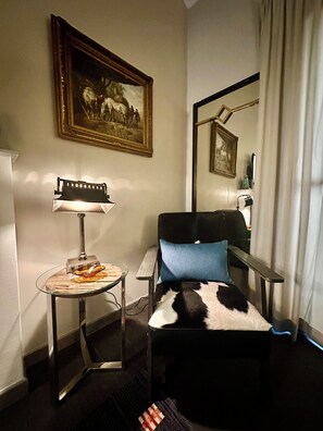A hotel room with a bed, a small table with a lamp, a painting on the wall, and a window with curtains.