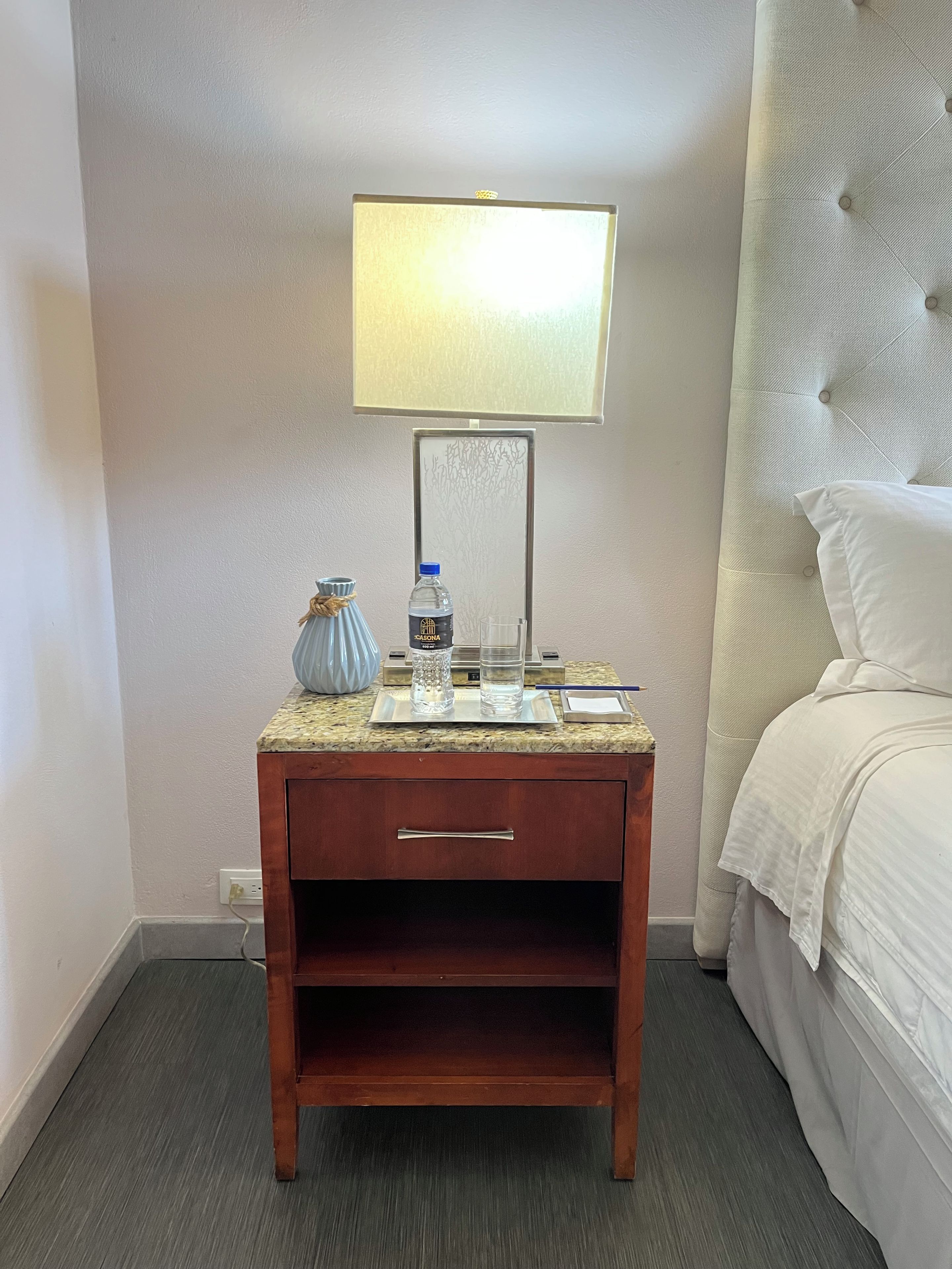 Habitación de hotel con cama, mesita de noche con lámpara, botella de agua, vaso y jarra.