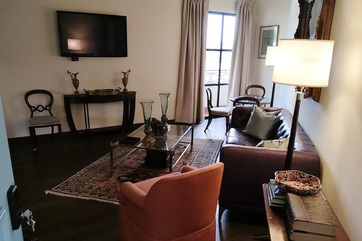 A living room with a sofa, armchair, TV, and a table with books and a lamp.