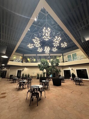 Un espacio interior amplio con varias mesas y sillas, un techo alto con iluminación decorativa y un arreglo floral central.