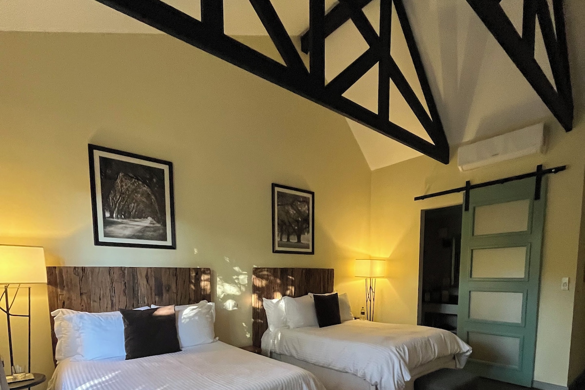 A hotel room with two beds, a bench, a patterned rug, and a wooden ceiling structure.