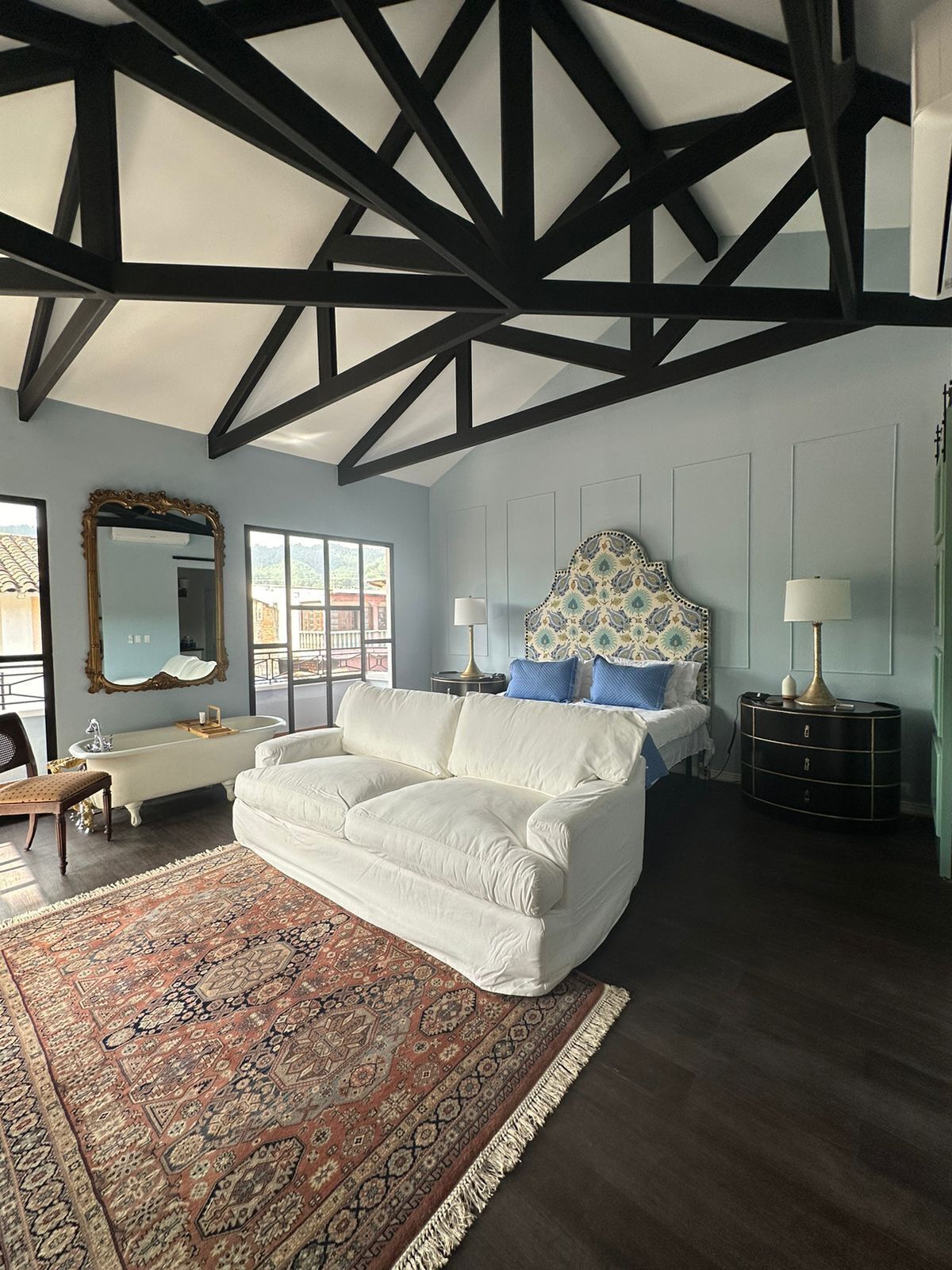 A bedroom with a large white sofa, a patterned headboard, a round black side table, and a classic white bathtub.