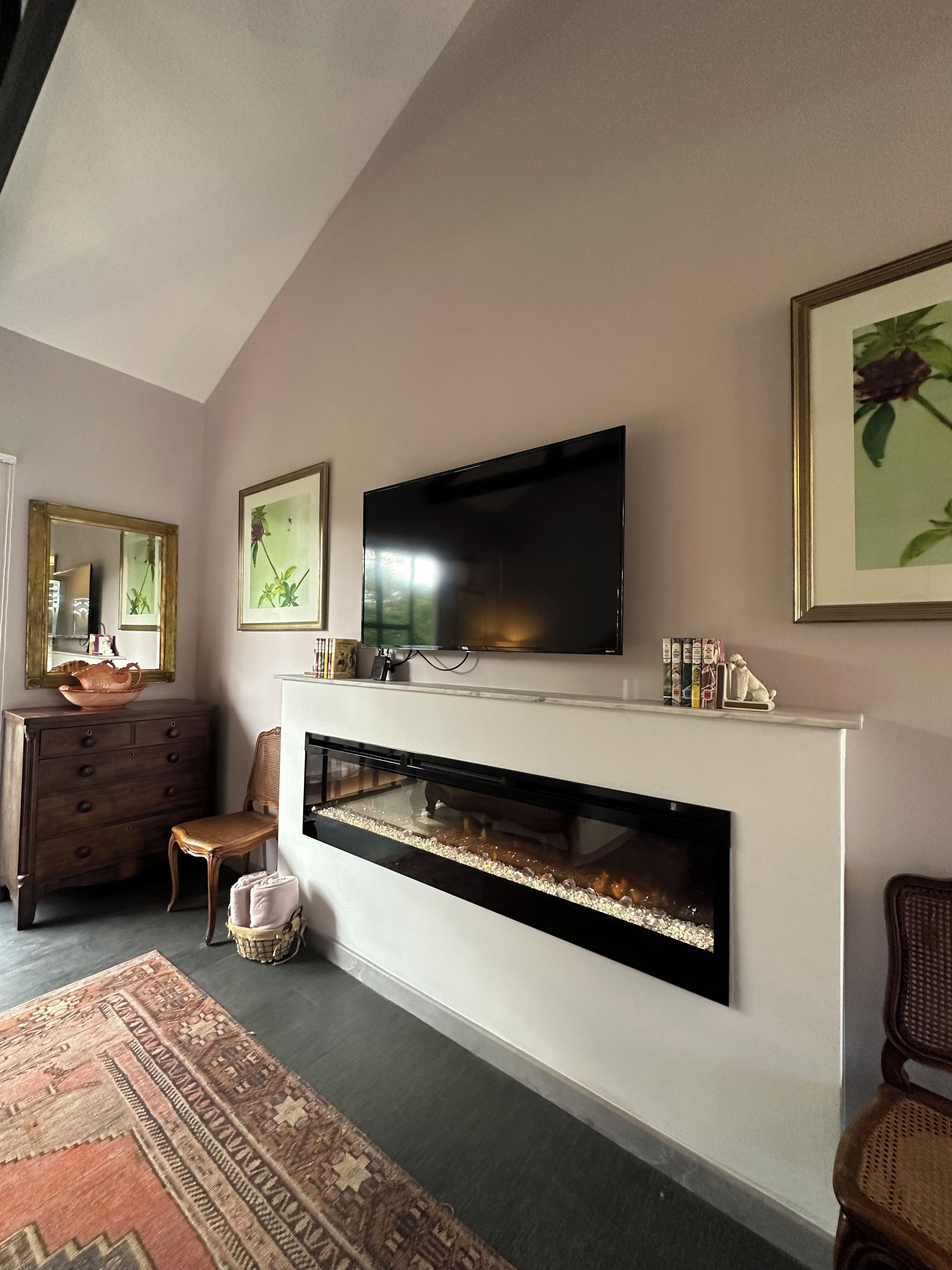 Una sala de estar con una televisión de pantalla plana sobre una chimenea, un aparador de madera, un sillón de mimbre y un tapete estampado.