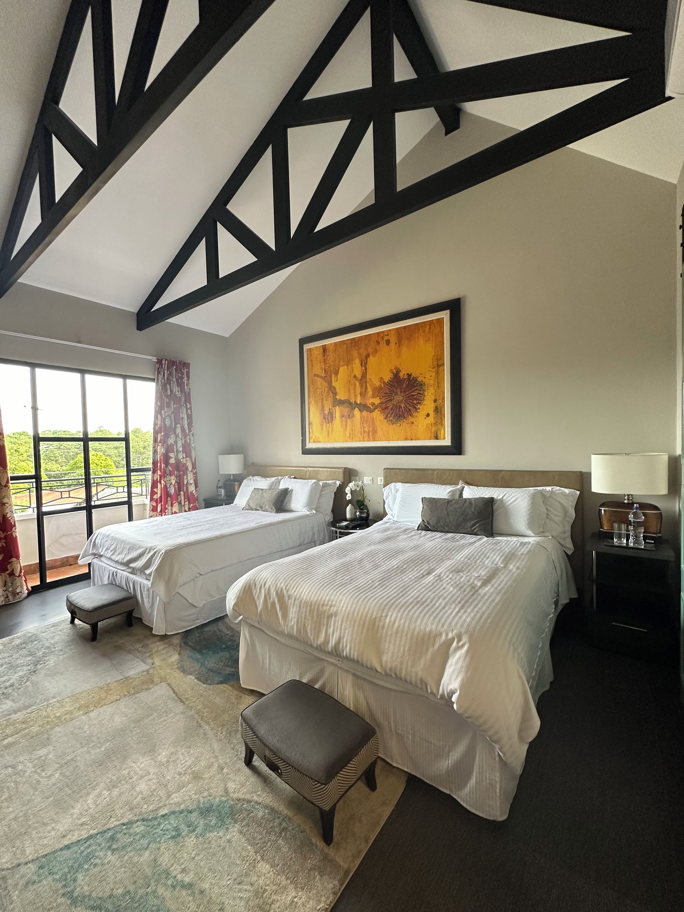 A hotel room with two beds, a large window, a painting, and a nightstand with a lamp.
