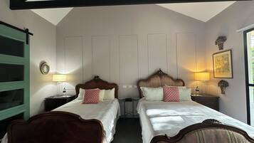 A room with two beds, a wooden ceiling, a window with a view, and a wall-mounted lamp.