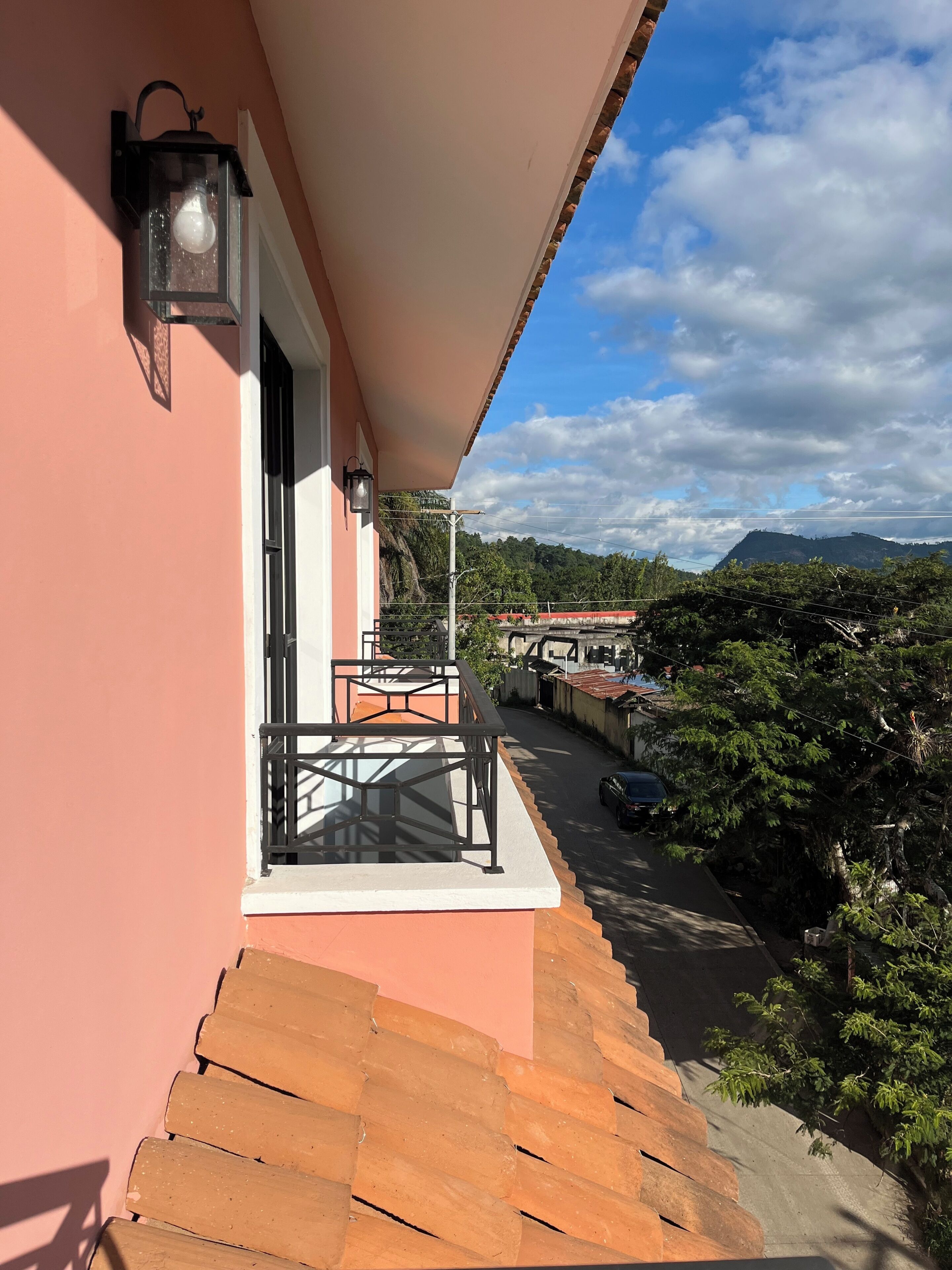 A balcony with a view of a road and greenery, a window with a metal railing, and a wall-mounted light fixture.